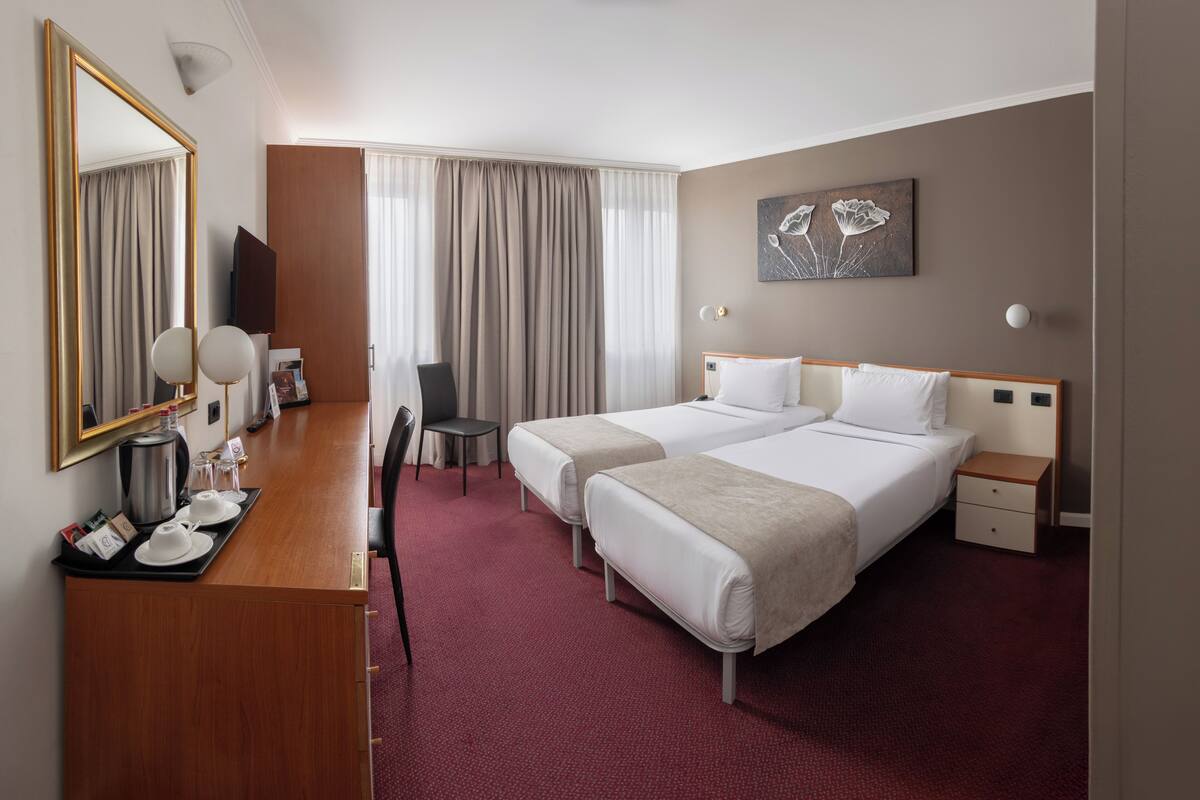 A hotel room with two beds, a desk, a chair, a coffee station, and a painting on the wall.