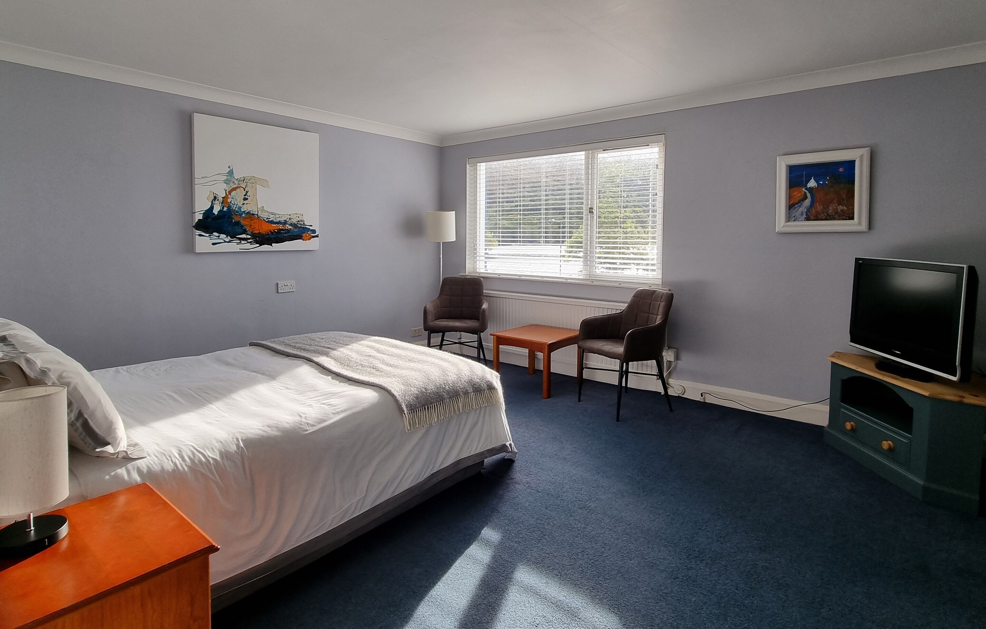 A hotel room with a bed, a desk, a chair, a TV, and artwork on the walls.