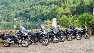 A row of parked motorcycles along a gravel path with a lake and forested hills in the background.