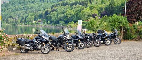 Une rangée de motos garées le long d’un chemin de gravier, avec un lac et des collines boisées en arrière-plan.