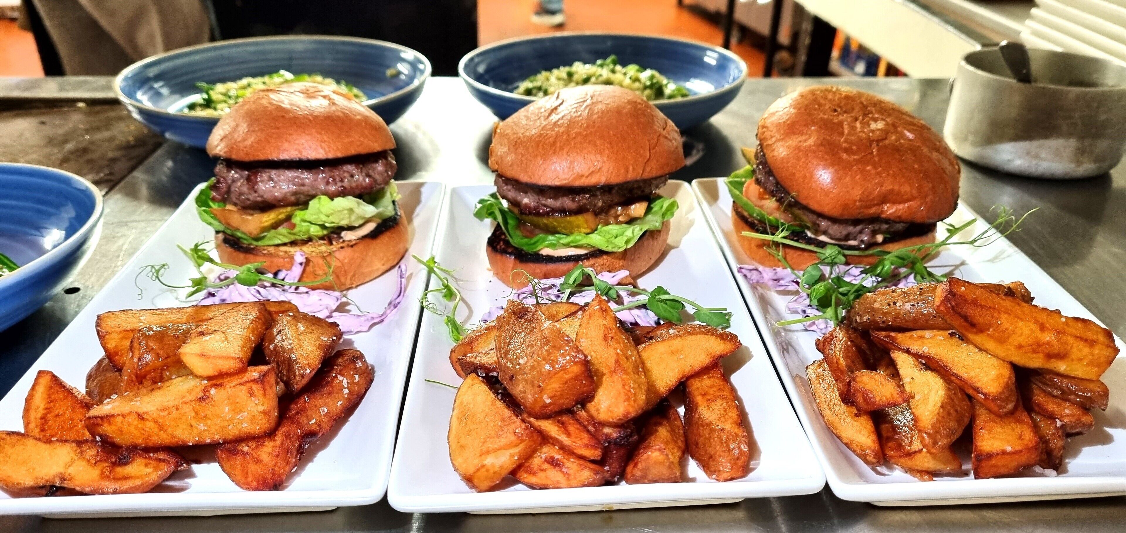 Tres hamburguesas con papas fritas en platos blancos, cada una acompañada de una ensalada en un tazón azul.