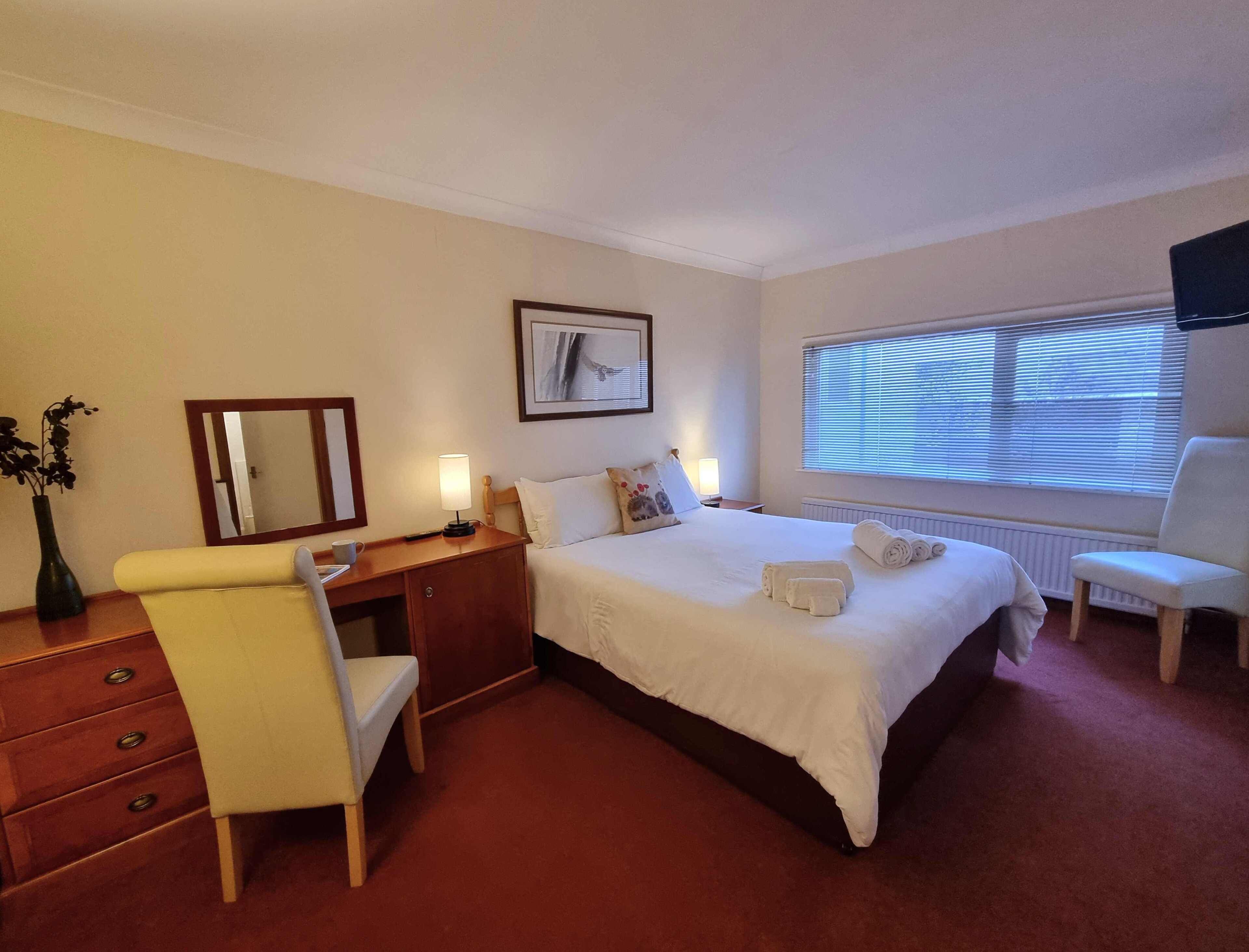 A hotel room with a bed, bedside table, chair, desk, and a window with blinds.