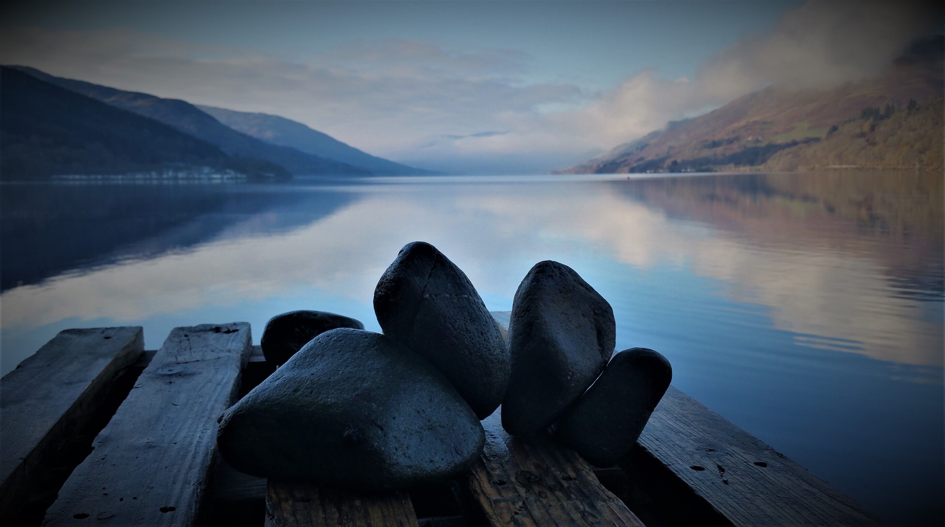 A wooden dock with large rocks in the foreground and a calm lake with mountains in the background.