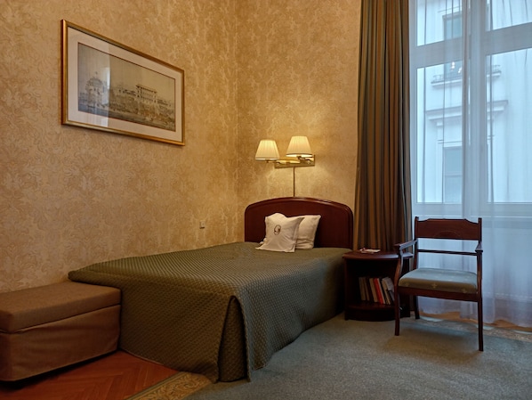 A hotel room with a bed, a nightstand with lamps, a chair, a small bookshelf, and a window with curtains.