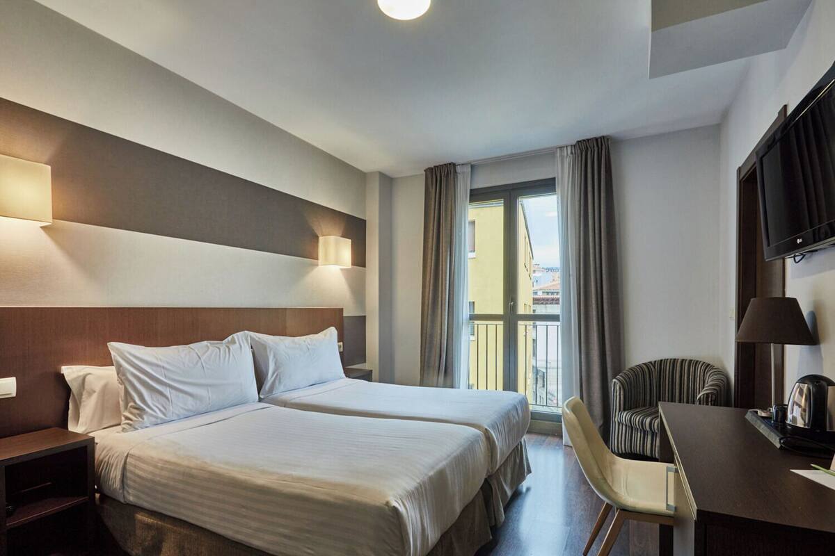 A hotel room with two beds, a desk, a chair, a TV, and a window with curtains.