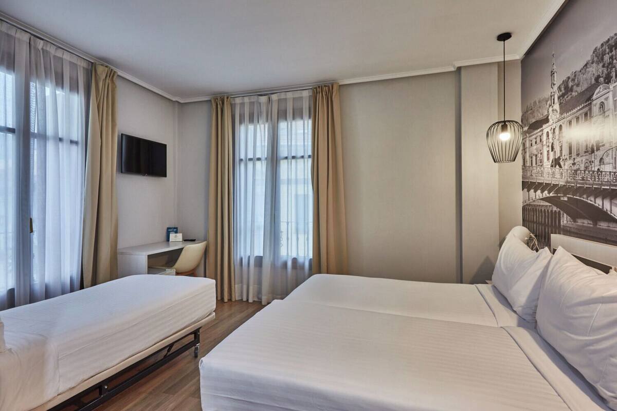 A hotel room with two beds, a desk, a chair, a TV, and a large window with curtains.