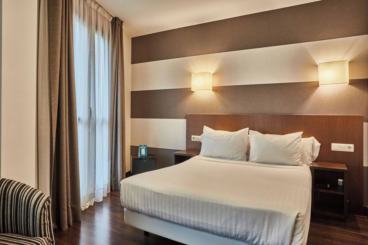 A hotel room with a large bed, two bedside tables, a striped chair, and a view of the outdoors through curtains.
