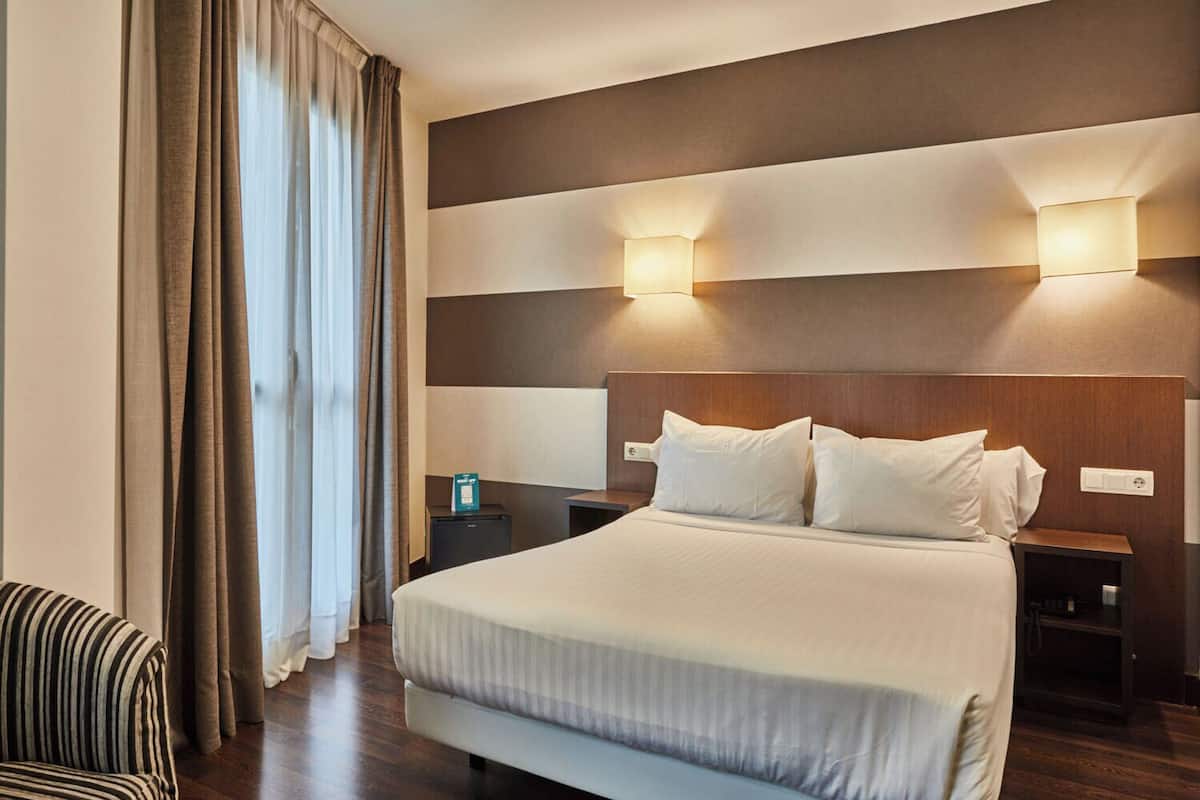 Quarto de hotel com uma cama grande, duas mesas de cabeceira, uma cadeira listrada e vista para o exterior através das cortinas.