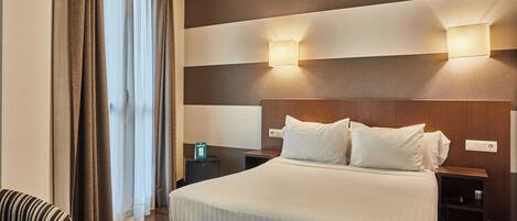 Habitación de hotel con una cama grande, dos mesitas de noche, una silla a rayas y vistas al exterior a través de cortinas.