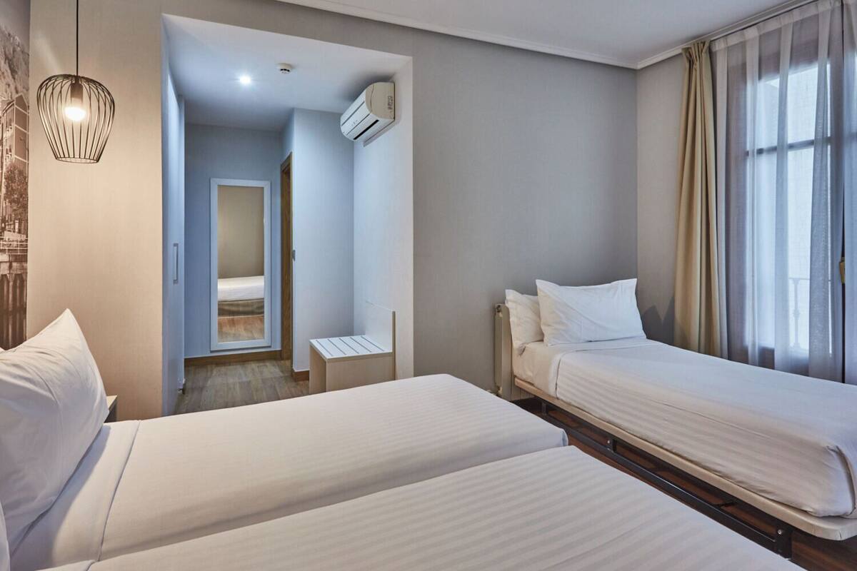 A hotel room with two beds, a small table, a chair, a ceiling light, an air conditioning unit, and a window with curtains.
