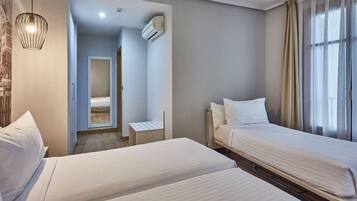 A hotel room with two beds, a small table, a chair, a ceiling light, an air conditioning unit, and a window with curtains.