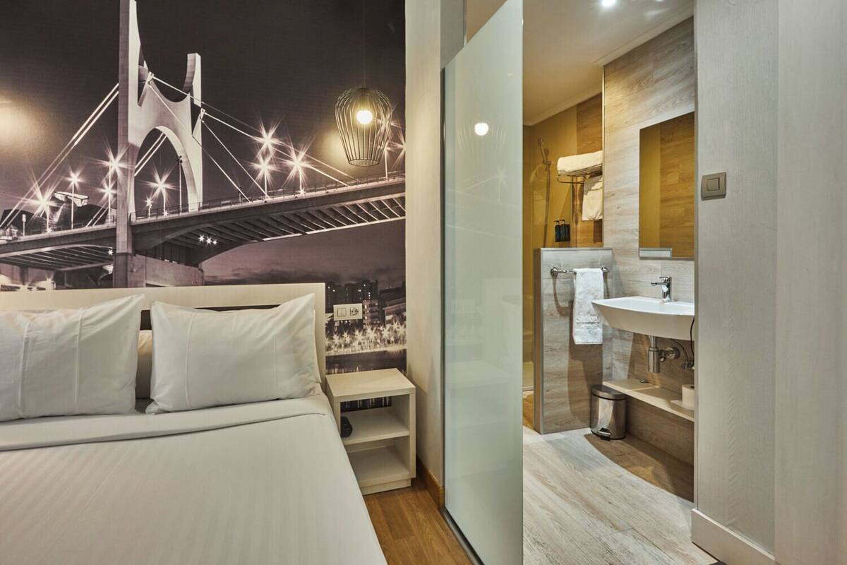 A modern hotel room with a bed, a nightstand, a glass partition, a bathroom with a sink and mirror, and a cityscape mural on the wall.