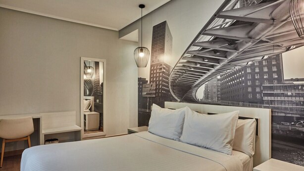 A modern hotel room with a bed, a desk, a chair, and a cityscape mural on the wall. - Silken Sirimiri (Bilbao)