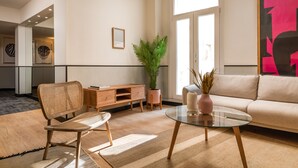 Un salon moderne avec une table basse en verre, un fauteuil en osier, un canapé beige, une table d’appoint en bois et une plante en pot.
