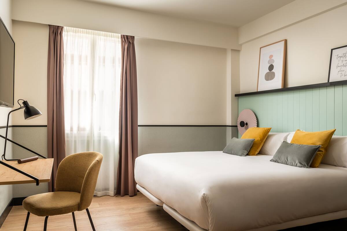 A bedroom with a bed, a desk with a chair, a lamp, and a window with curtains.