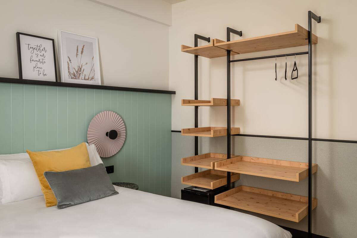 A bedroom with a bed, pillows, a wall-mounted shelf, and two framed pictures.