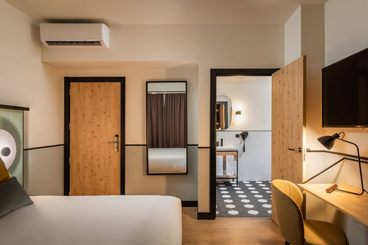 A modern hotel room with a bed, a desk, and a bathroom visible through an open door.