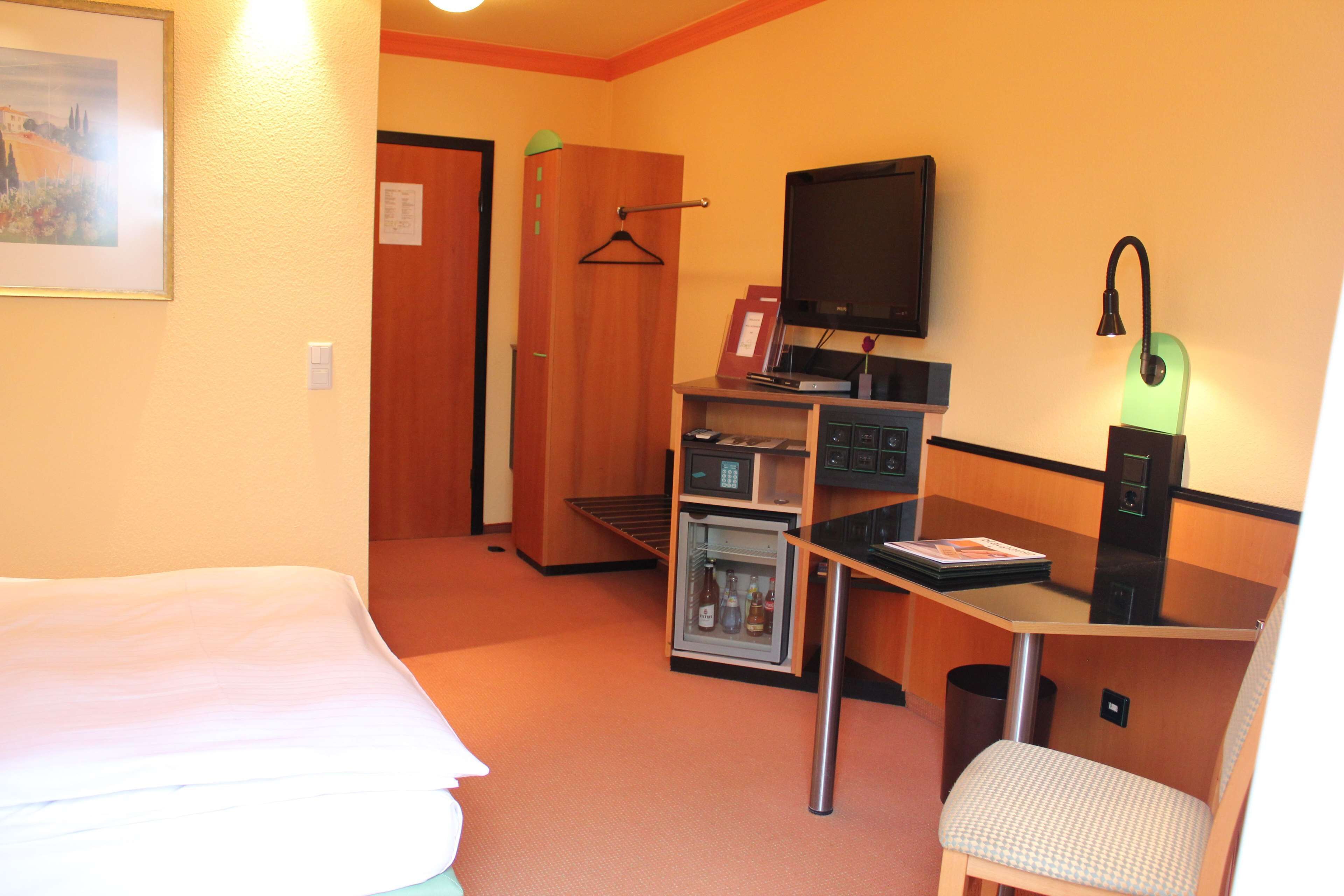 Habitación de hotel con cama, escritorio con computadora, televisión, refrigerador pequeño y una silla.