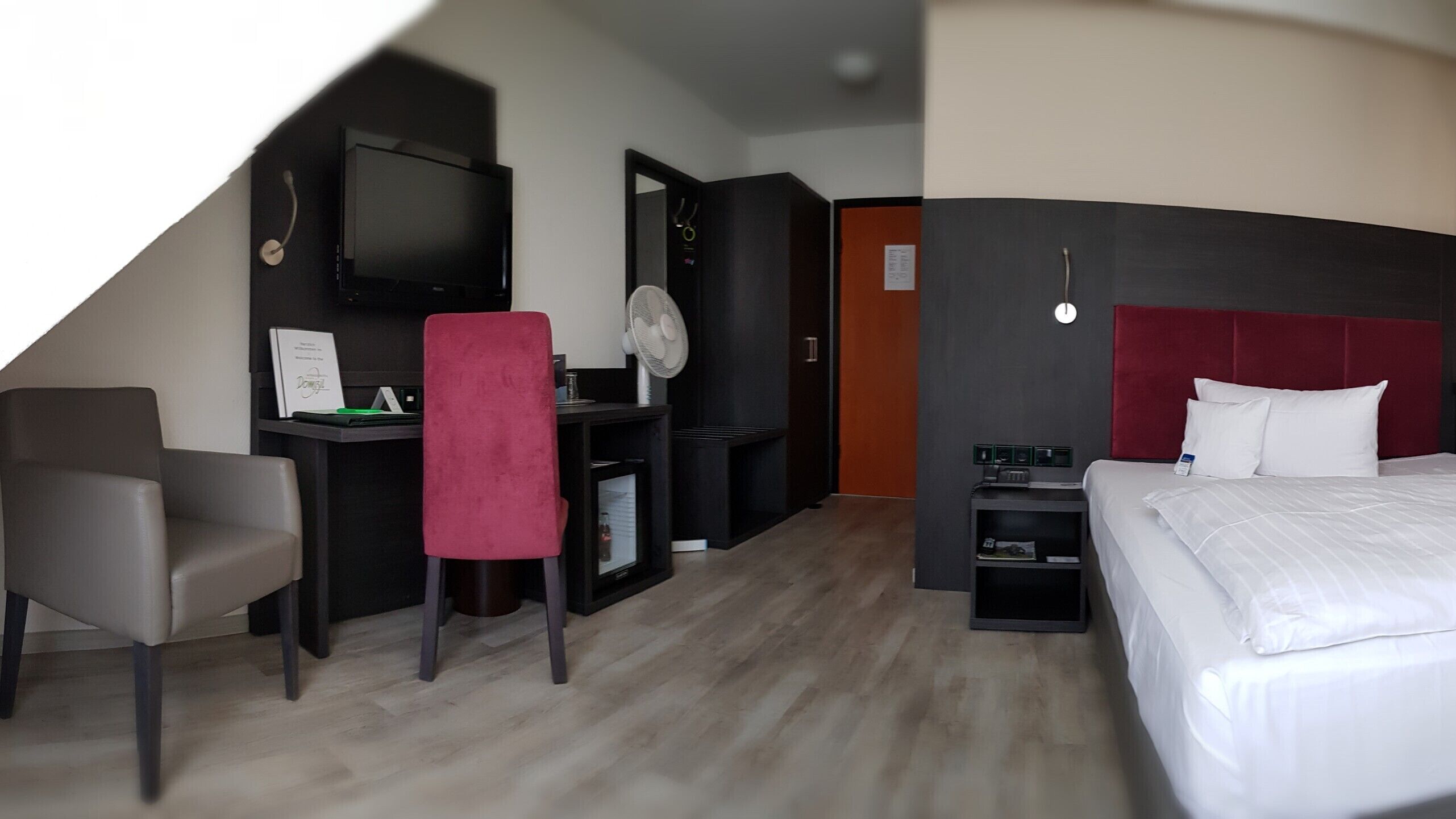 A hotel room with a bed, a desk with a chair, a TV, a fan, and a red accent chair.