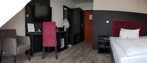 A hotel room with a bed, a desk with a chair, a TV, a fan, and a red accent chair.