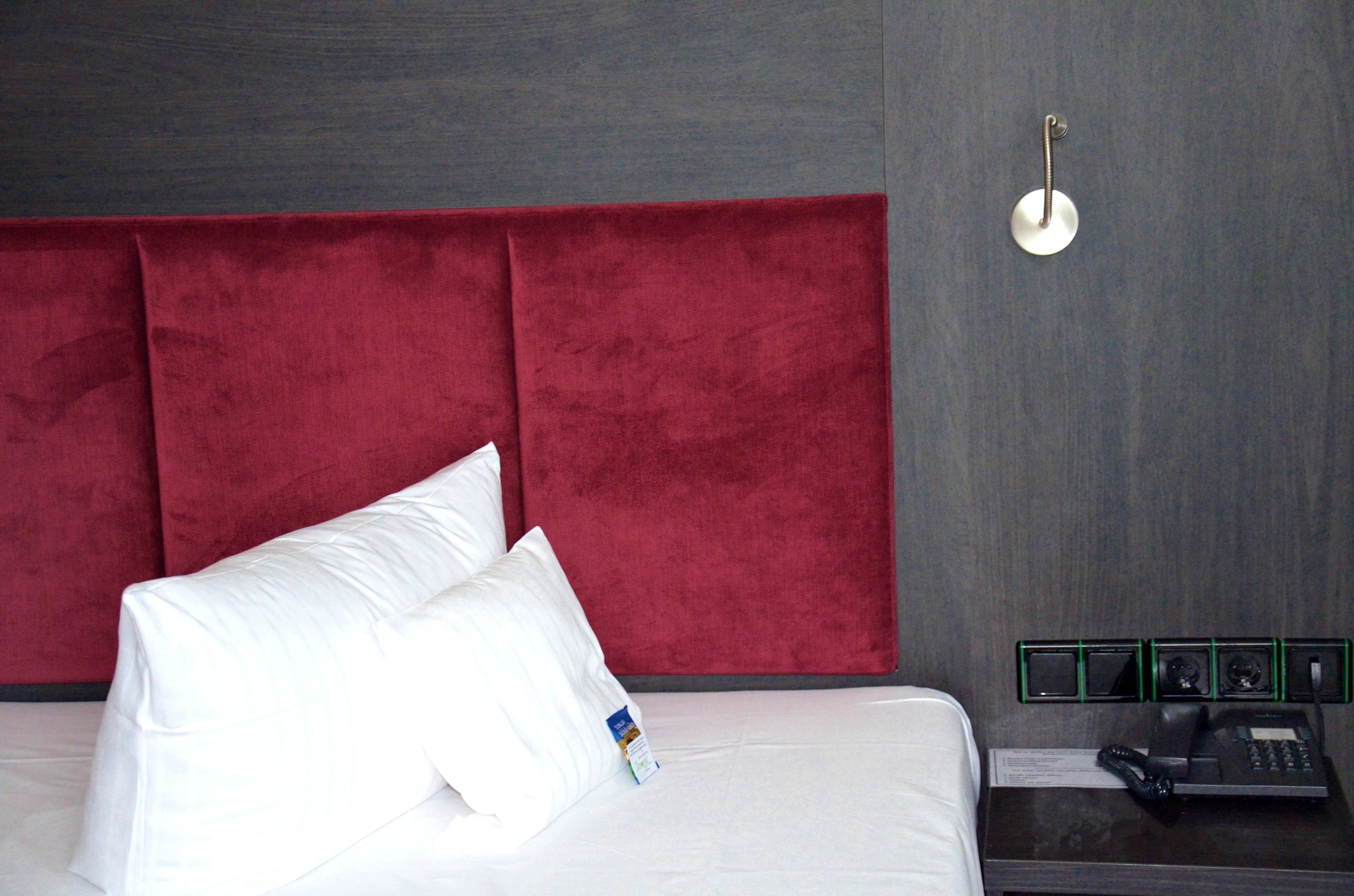 Habitación de hotel con un cabecero rojo grande, ropa de cama blanca, una mesita de noche con teléfono y lámpara, y una lámpara de pared.