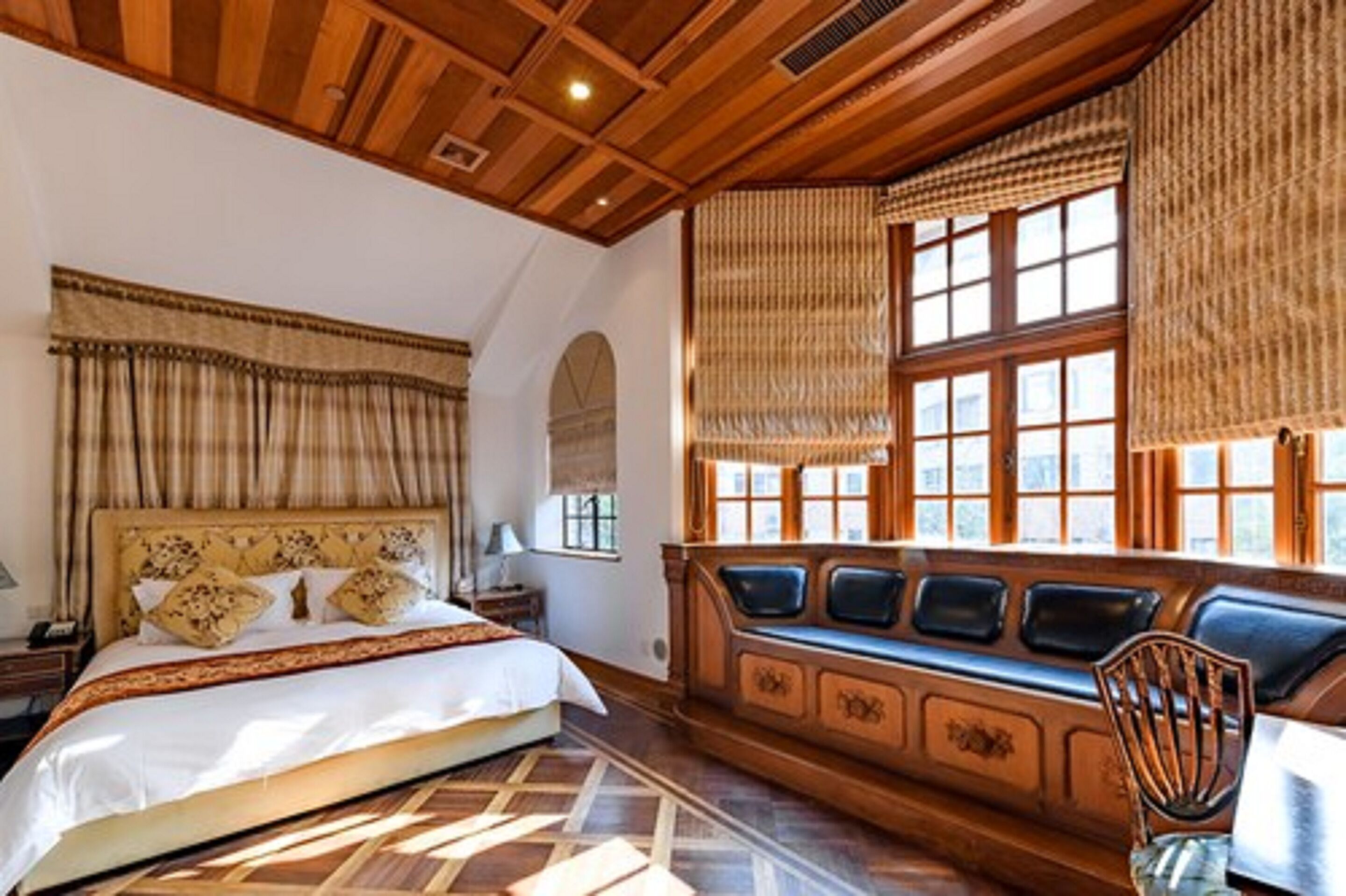 A bedroom with a large bed, a wooden bench with cushions, a window with blinds, and a patterned rug.
