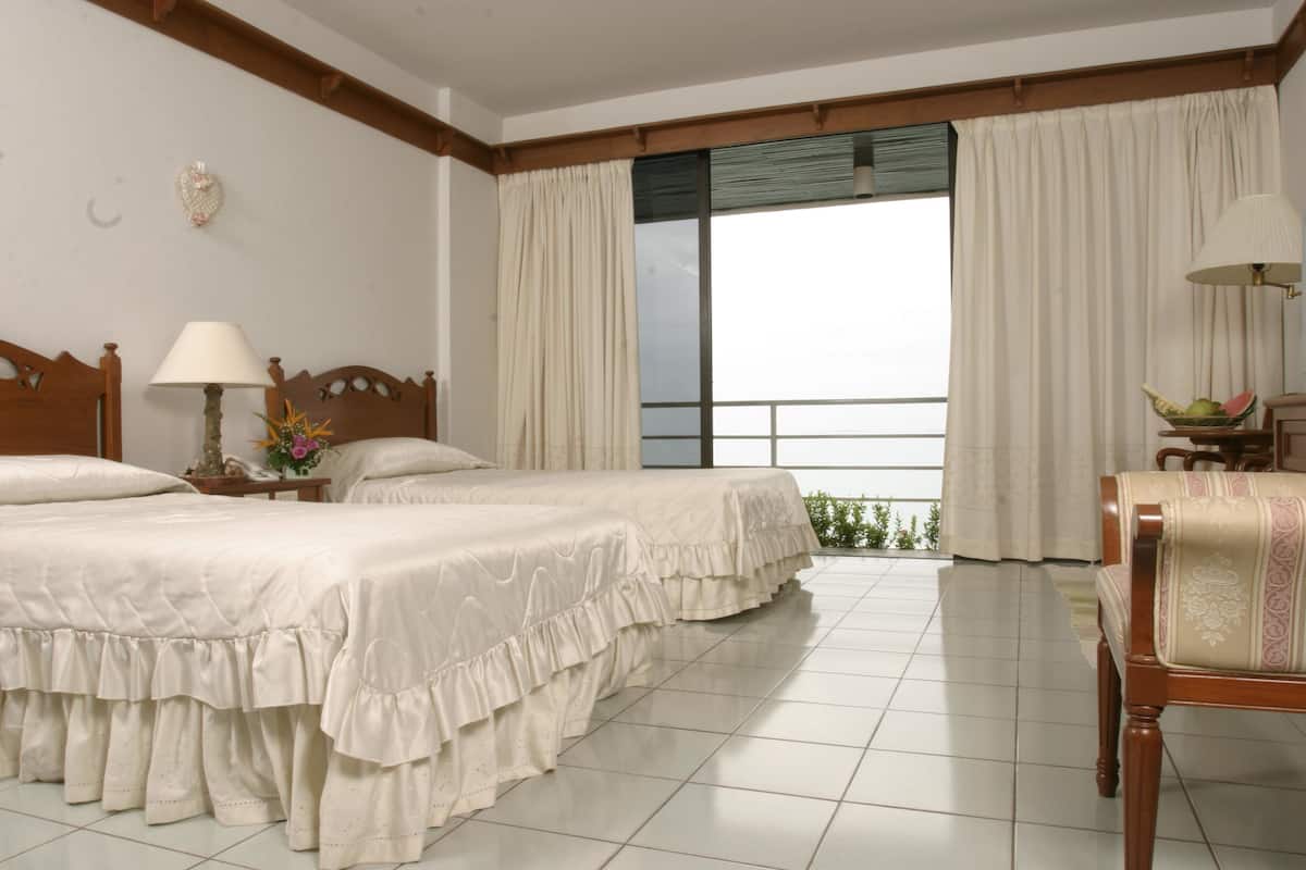 A hotel room with two beds, a balcony with curtains, a wooden chair, and a nightstand with a lamp.