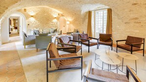 Une pièce spacieuse aux murs de pierre, dotée d’une cheminée et d’un coin salon confortable avec plusieurs fauteuils et tables.