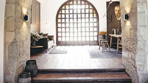 Un portail en arc donne accès à une pièce dotée d’une grande fenêtre, d’une table avec des chaises et d’un luminaire suspendu.