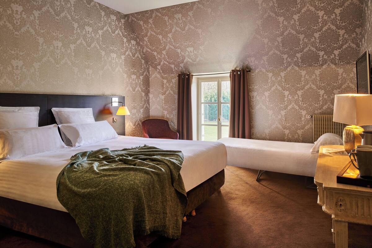 A hotel room with two beds, a nightstand, a lamp, and a window with curtains.