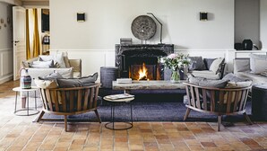 Un salon chaleureux avec une cheminée, deux fauteuils en bois, une petite table d’appoint et un canapé avec des coussins.