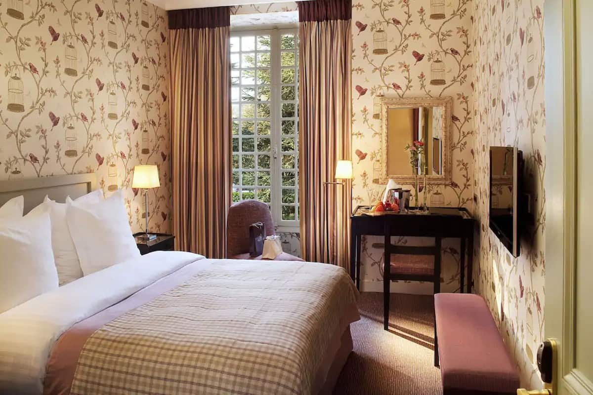 A hotel room with floral wallpaper, a bed with white linens, two bedside lamps, a desk with a mirror, and a bench.