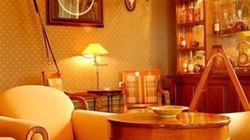 A room with a yellow patterned wall, a wooden table with a lamp, a framed picture, a stuffed animal, and a shelf with various items.