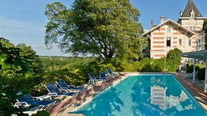 A swimming pool with lounge chairs, surrounded by greenery and a large building with a tower.