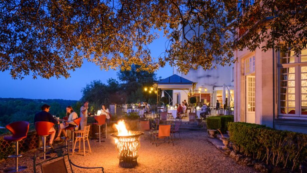 An outdoor dining area with a fire feature, tables, chairs, and a building in the background.