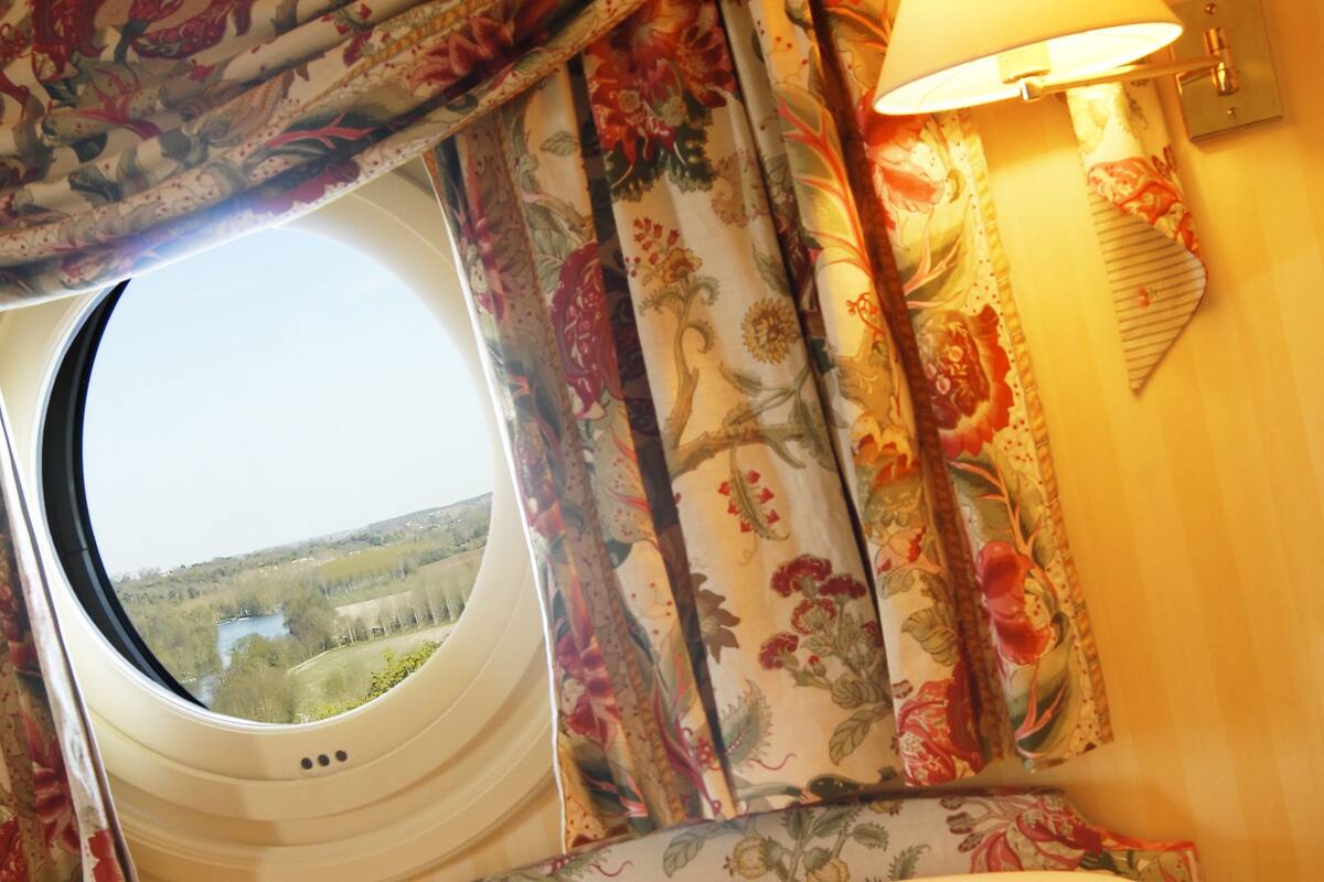 A bedroom with a window, floral curtains, a bed with white pillows, and a wall-mounted lamp.