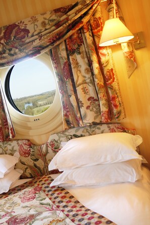 A bedroom with a window, floral curtains, a bed with white pillows, and a wall-mounted lamp.