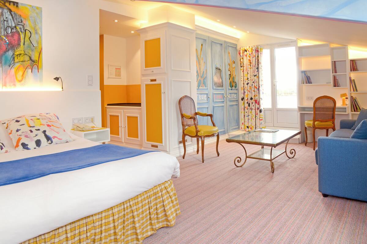 A hotel room with a bed, a desk, a chair, a sofa, and a bookshelf.