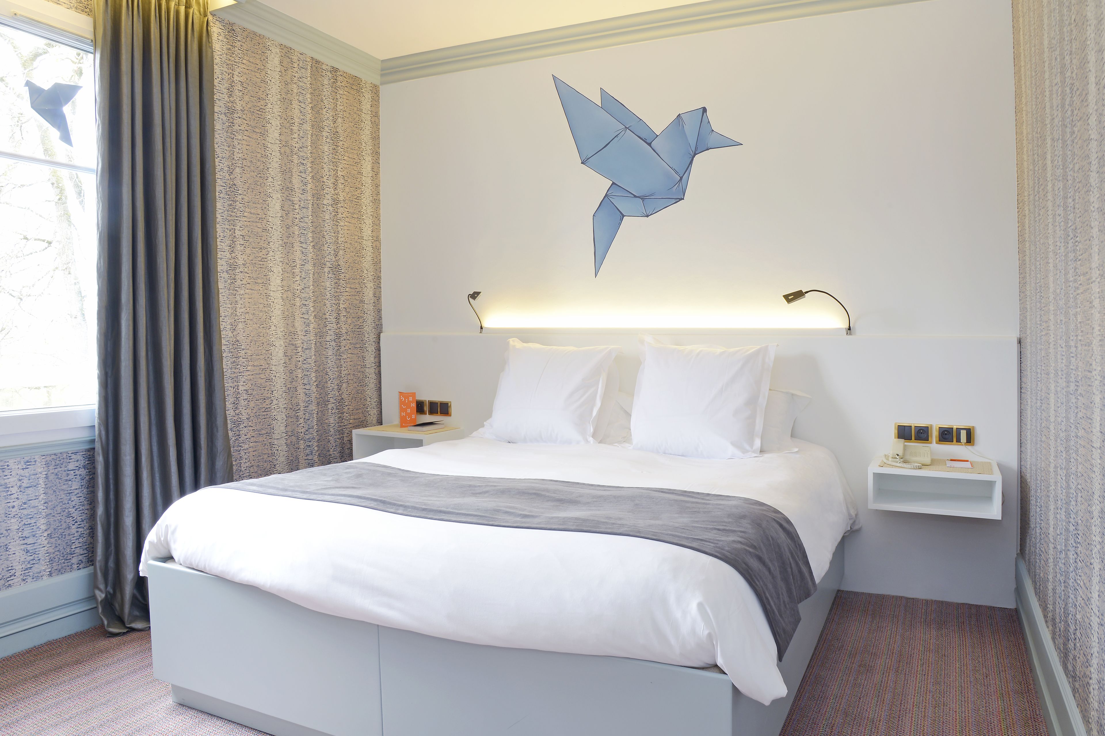 A modern hotel room with a large bed, a bedside table, a wall-mounted lamp, and a decorative wall piece.