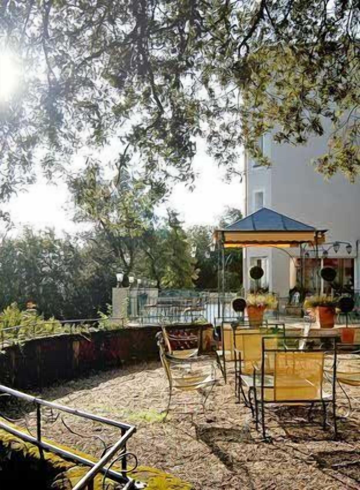 An outdoor seating area with tables and chairs, surrounded by greenery and a building in the background.