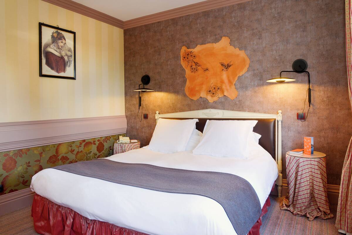 A neatly made bed with white linens and a gray throw, a framed picture on the wall, and two wall-mounted lamps.