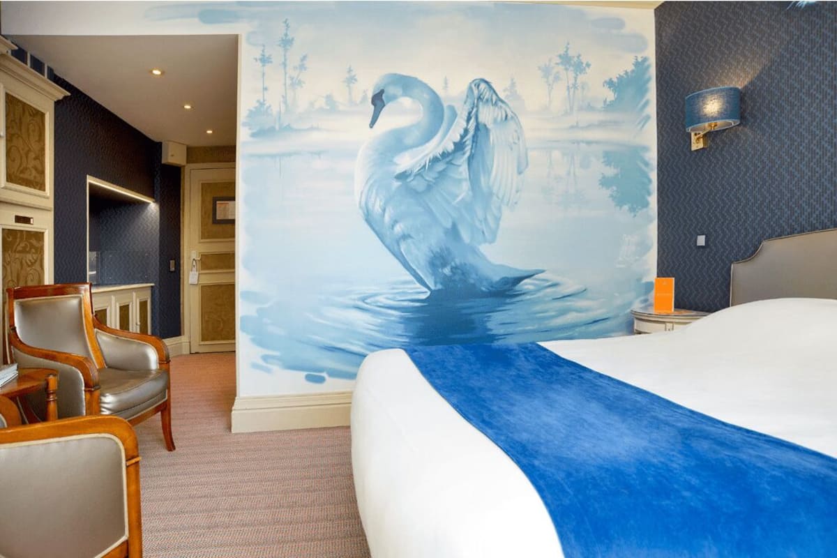 A hotel room with a large mural of a swan on a lake, a bed with a blue accent, a wooden chair, and a nightstand with a lamp.
