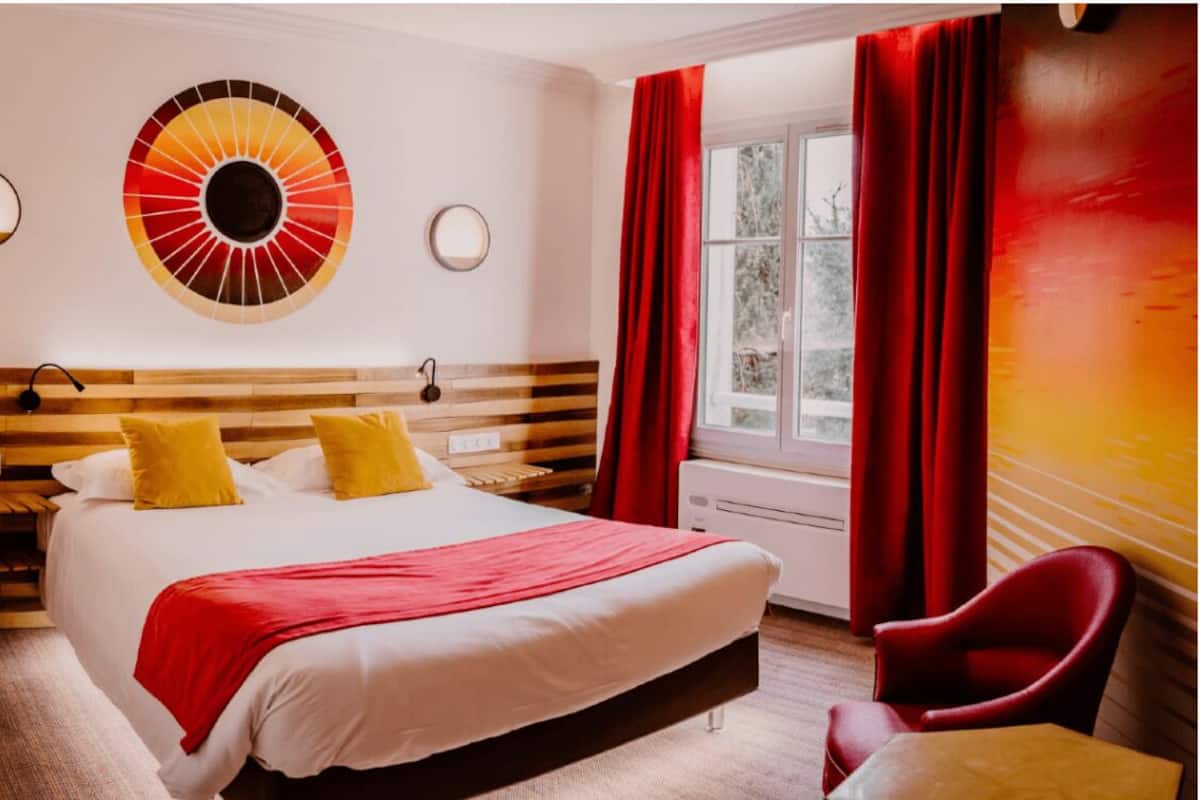 A hotel room with a large bed, two bedside tables, a red and yellow wall decoration, a window with red curtains, and a red chair.