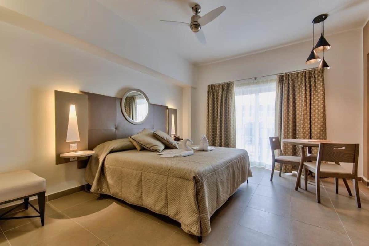 A hotel room with a bed, bedside table, lamp, mirror, and a dining area with chairs and a table.