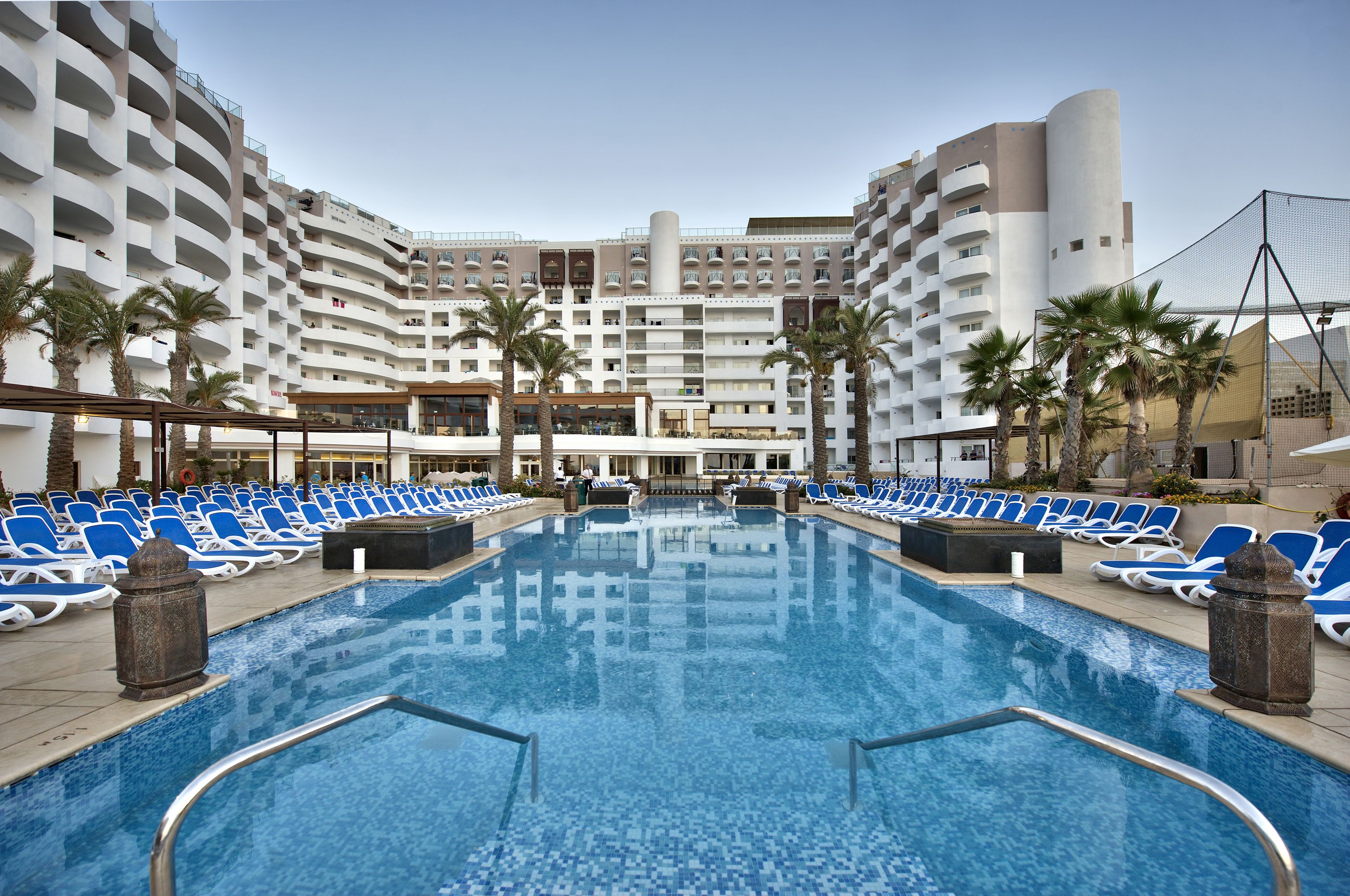 A hotel pool area with multiple levels of balconies, palm trees, and sun loungers.
