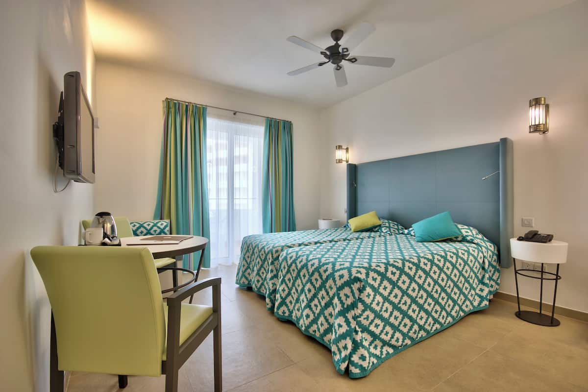 A hotel room with a bed, a desk with a chair, a ceiling fan, and a bedside table with a lamp.