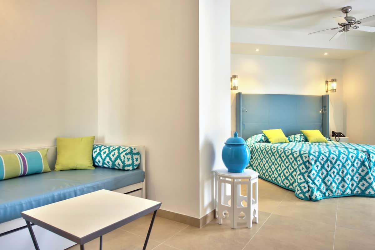 A room with a sofa, a bed with patterned bedding, a small white table, and a blue vase on a white stool.
