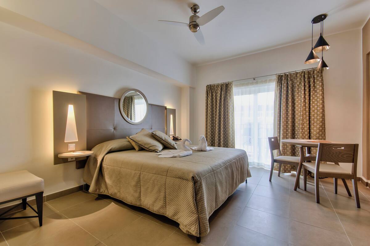 A hotel room with a bed, bedside table, lamp, mirror, and a dining area with a table and chairs.