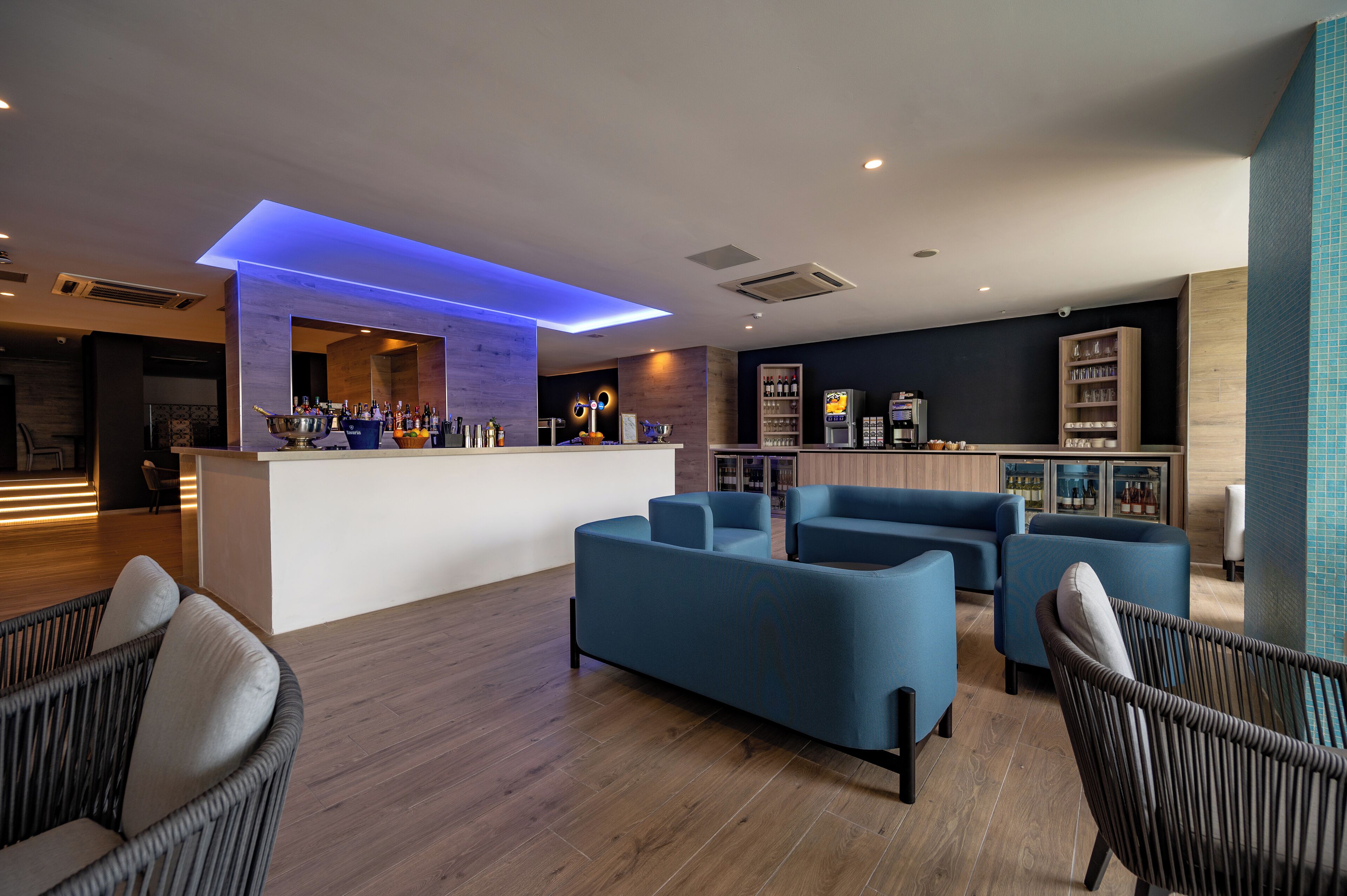 Un área de estar moderna con un bar, sofás azules, sillas de mimbre y suelo de madera.