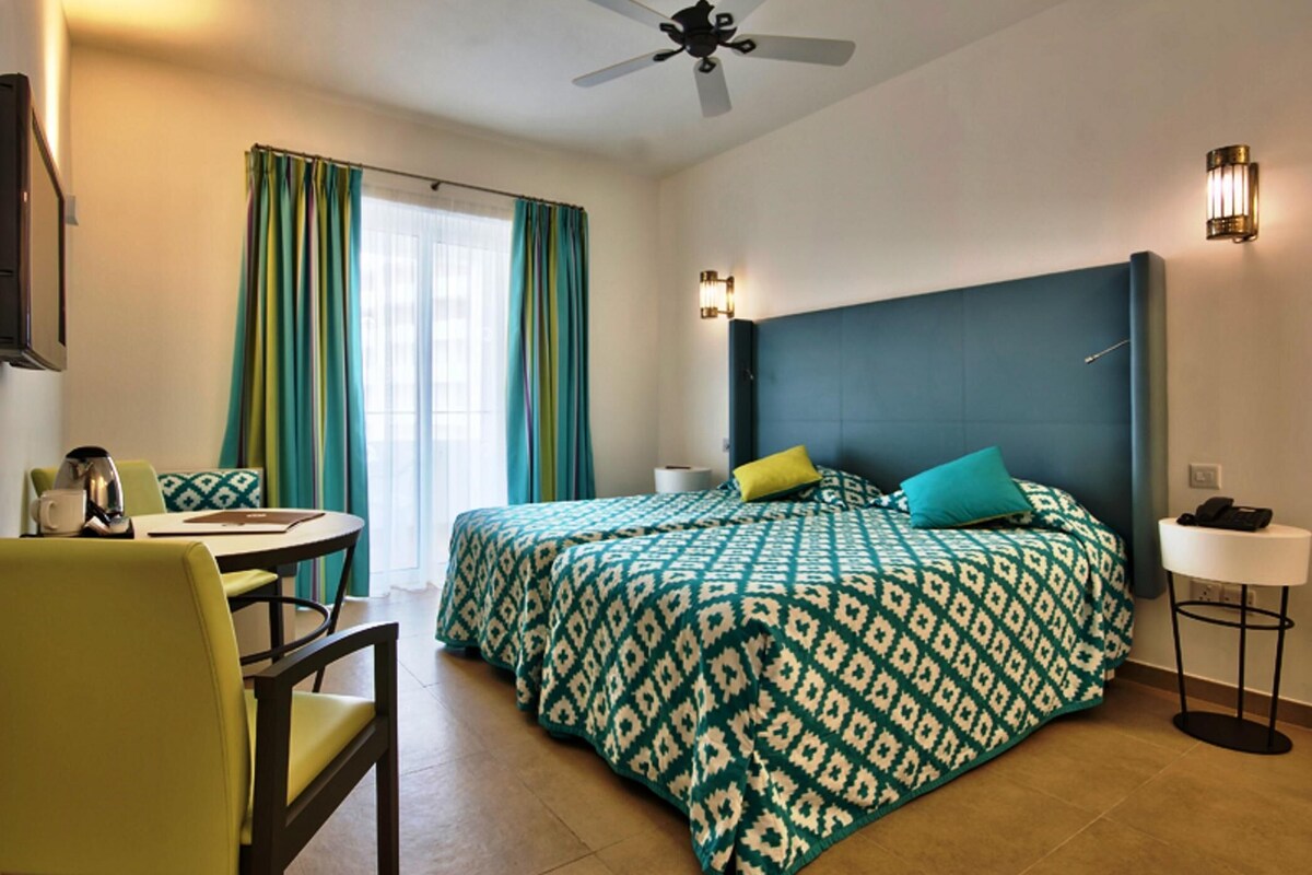 A hotel room with a bed, a desk, a chair, a ceiling fan, and a window with curtains.